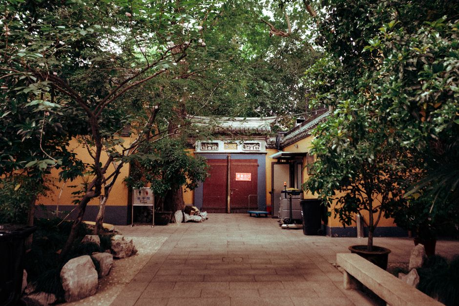 A tranquil courtyard featuring a paved stone walkway leading to a traditional Asian-style gate with a curved tiled roof, surrounded by lush, green trees and bushes. The garden beds on either side of t