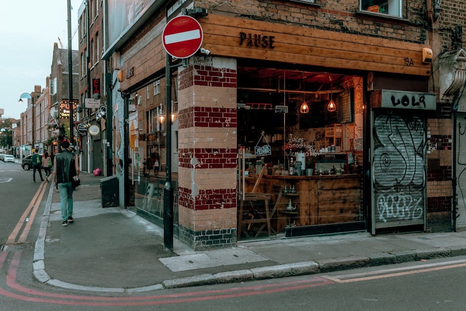 An urban street corner featuring a small café with a wooden sign that reads 'PULSE' above the entrance, located at 134 Shoreditch. The café has large glass windows revealing a warm, cozy interior il